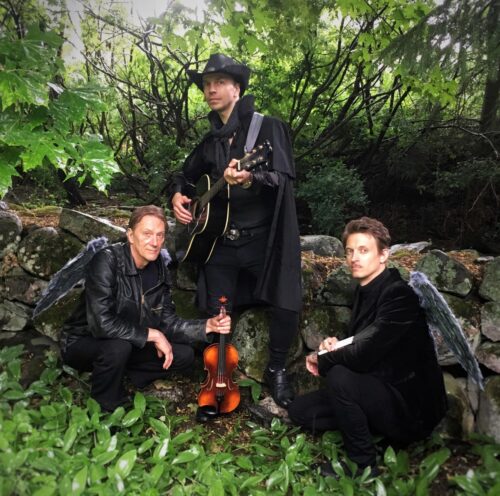 Tomi Pulkkinen ja trio ulkona, takana puu. Triolla on soittimet käsissään. Tomi Pulkkinen med sin trio. En träd in bakgrunden, hela trio har sina instrumenter.